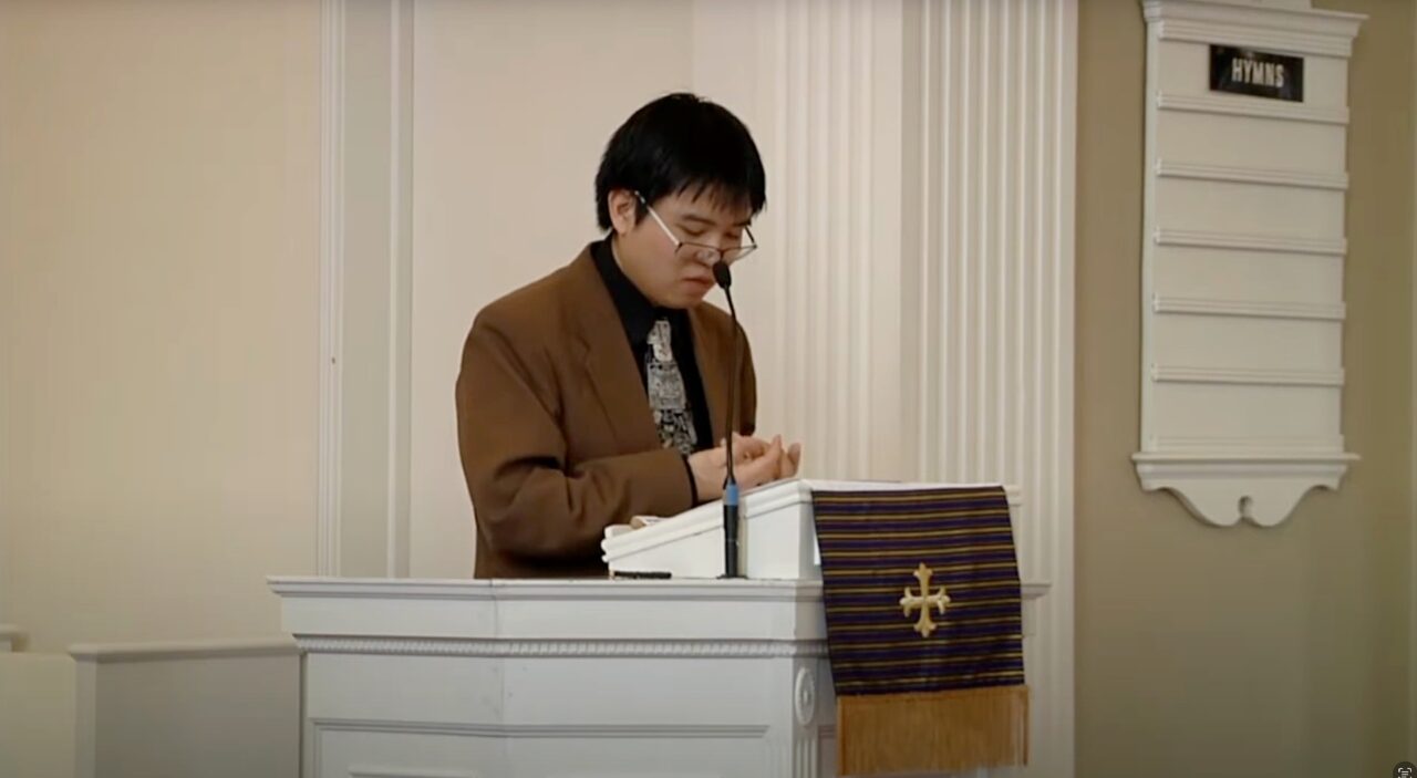 Asian American professor wearing blue blazer and navy shirt lectures behind white pulpit in chapel