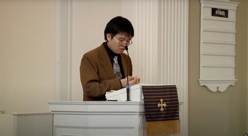 Asian American professor wearing blue blazer and navy shirt lectures behind white pulpit in chapel