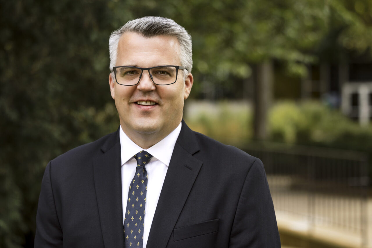 Professor wearing glasses, a dark suit and blue tie smiles at the camera