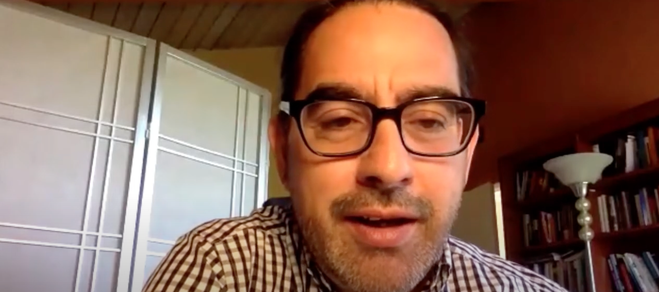 Man in glasses and checkered shirt looks at computer camera with a slight smile as he lectures to online audience