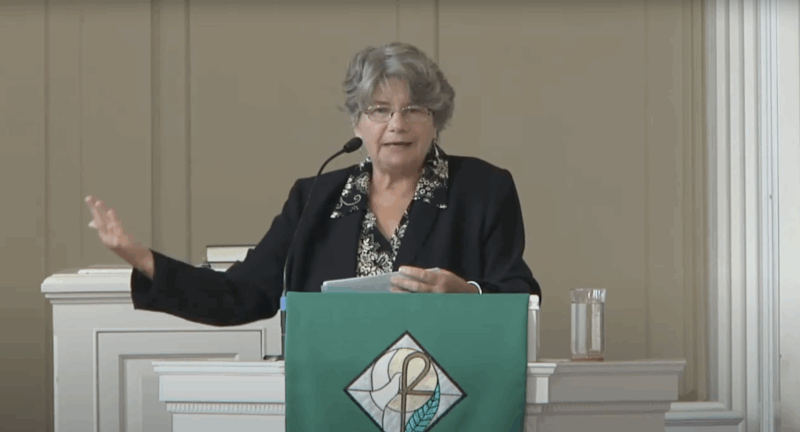 Gray-haired white female professor lectures from behind the pulpit in a chapel.