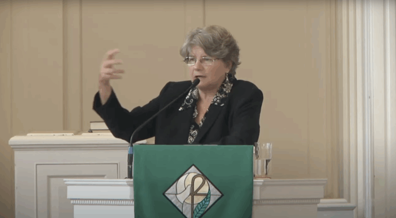 Gray-haired white female professor lectures from behind the pulpit in a chapel.