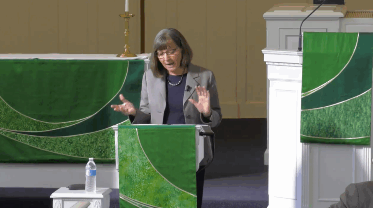 gray-haired females professor in gray blazer lectures from behind lectern draped in liturgical cloth