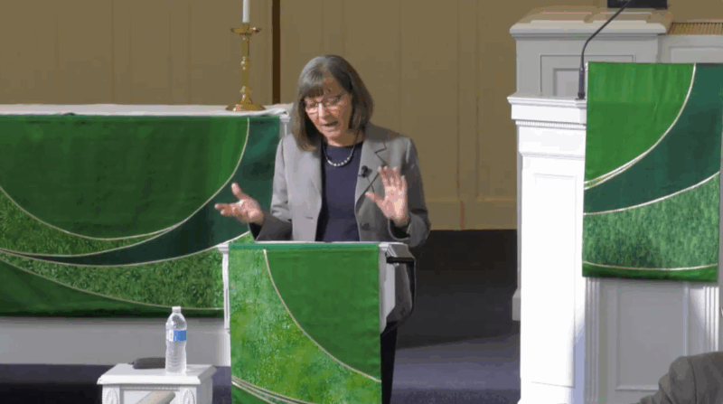 gray-haired females professor in gray blazer lectures from behind lectern draped in liturgical cloth