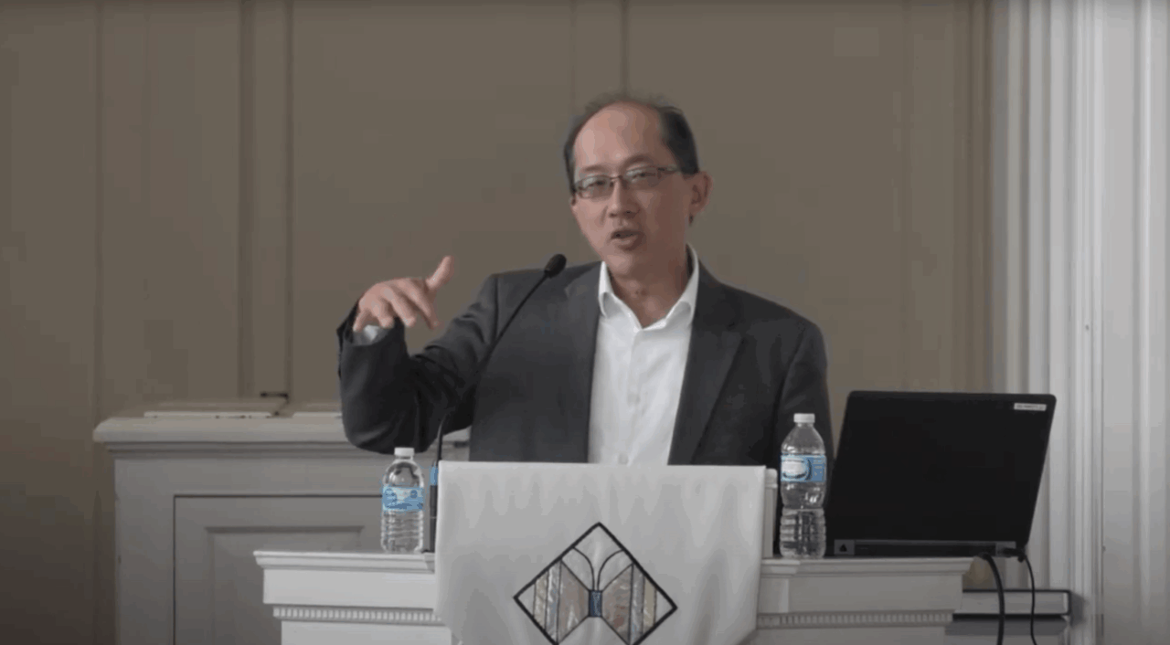 Malaysian-American lecturer in white shirt and gray blazer lectures from behind a pulpit in a chapel with laptop and bottle of water