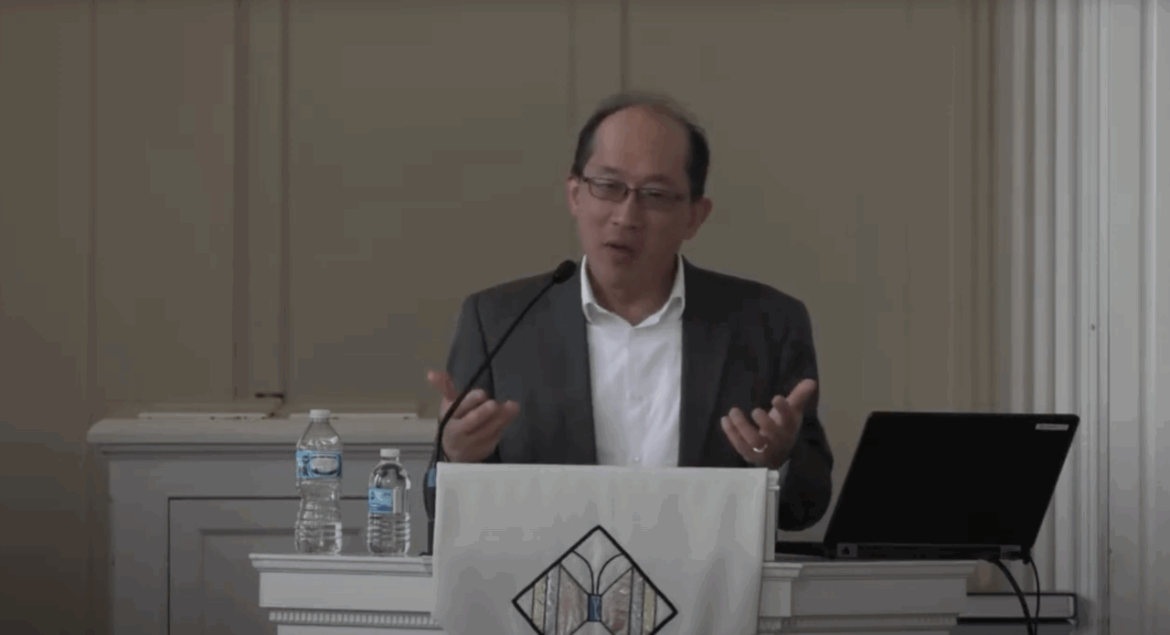 Malaysian-American lecturer in white shirt and gray blazer lectures from behind a pulpit in a chapel with laptop and bottle of water
