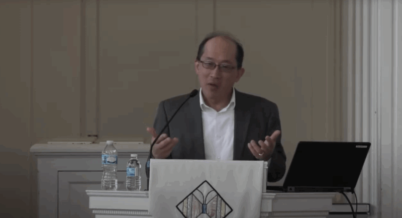 Malaysian-American lecturer in white shirt and gray blazer lectures from behind a pulpit in a chapel with laptop and bottle of water