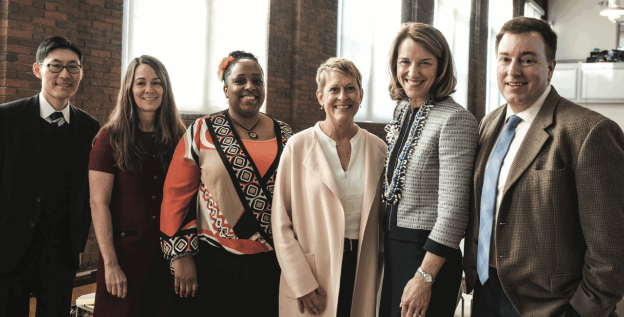 Diverse group of scholars stands in front of brick wall smiling at the camera