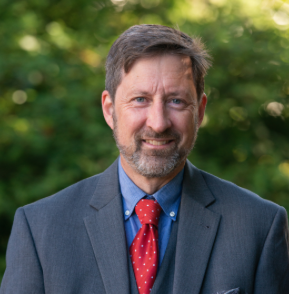 White dark-haired professor in gray jacket and red tie smiles at the camera in front of greenery