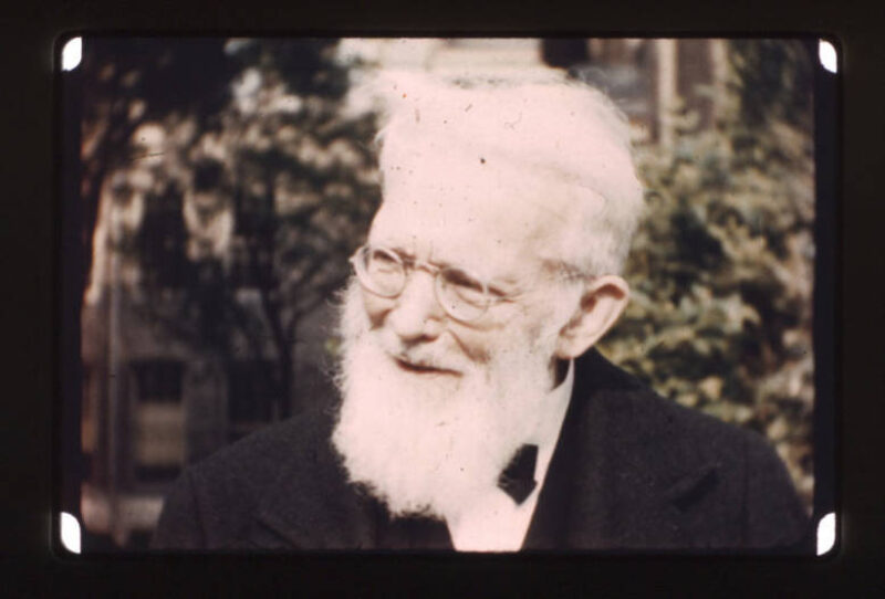 Elderly white professor with white hair, beard, and round spectacles smiles as he engages in conversation