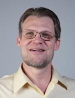 White man with glasses and beard wearing a light yellow collared shirt smiles at the camera