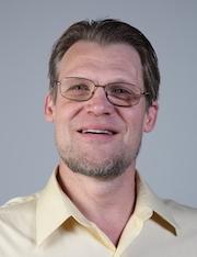 White man with glasses and beard wearing a light yellow collared shirt smiles at the camera