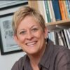 Professor with short blond hair and brown shirt smiles at the camera in front of bookshelf in office