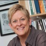 Professor with short blond hair and brown shirt smiles at the camera in front of bookshelf in office