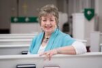 Gray-haired woman wearing blue scarf sits in chapel pew and smiles at the camera