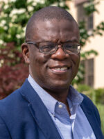 Bald black professor with glasses and navy jacket smiles at the camera