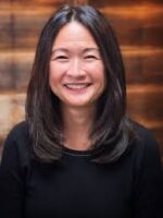 Asian-American woman with medium-length hair smiles at the camera in front of brick wall