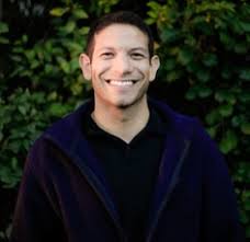 Latino man wearing black shirt and standing against a background of greenery smiles at the camera