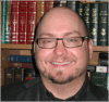 Bald white man with black-rimmed glasses sits in front of books and smiles at camera