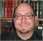Bald white man with black-rimmed glasses sits in front of books and smiles at camera