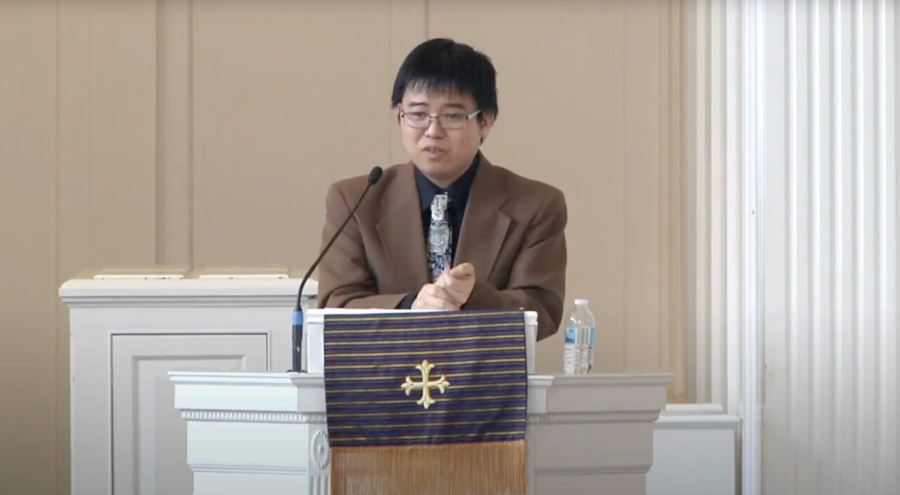 Asian American professor in navy shirt and brown blazer lectures at a pulpit
