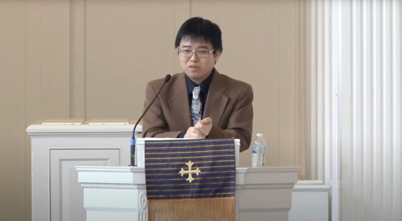 Asian American professor in navy shirt and brown blazer lectures at a pulpit