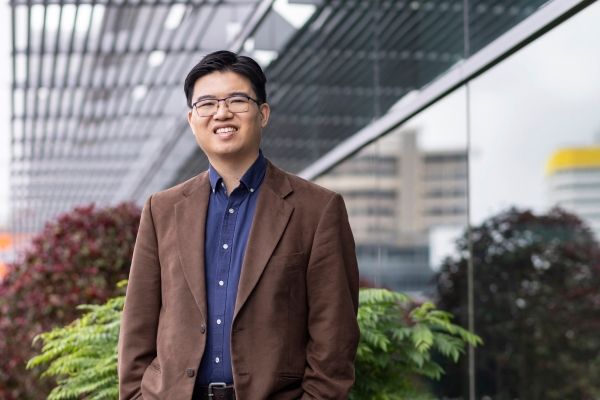 Asian-American man wearing glasses and brown blazer smiles at the camera