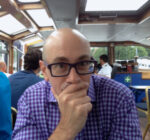 Bald white man in black-rimmed glasses and purple checkered shirt looks at the camera