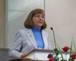 White woman wearing blue turtleneck and gray blazer speaks behind a podium