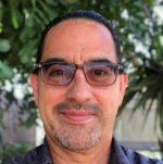 Latino man with glasses and graying beard smiles at the camera