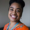 Black woman with orange shirt and blue necklace smiles at the camera