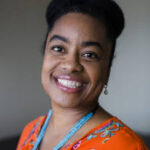 Black woman with orange shirt and blue necklace smiles at the camera