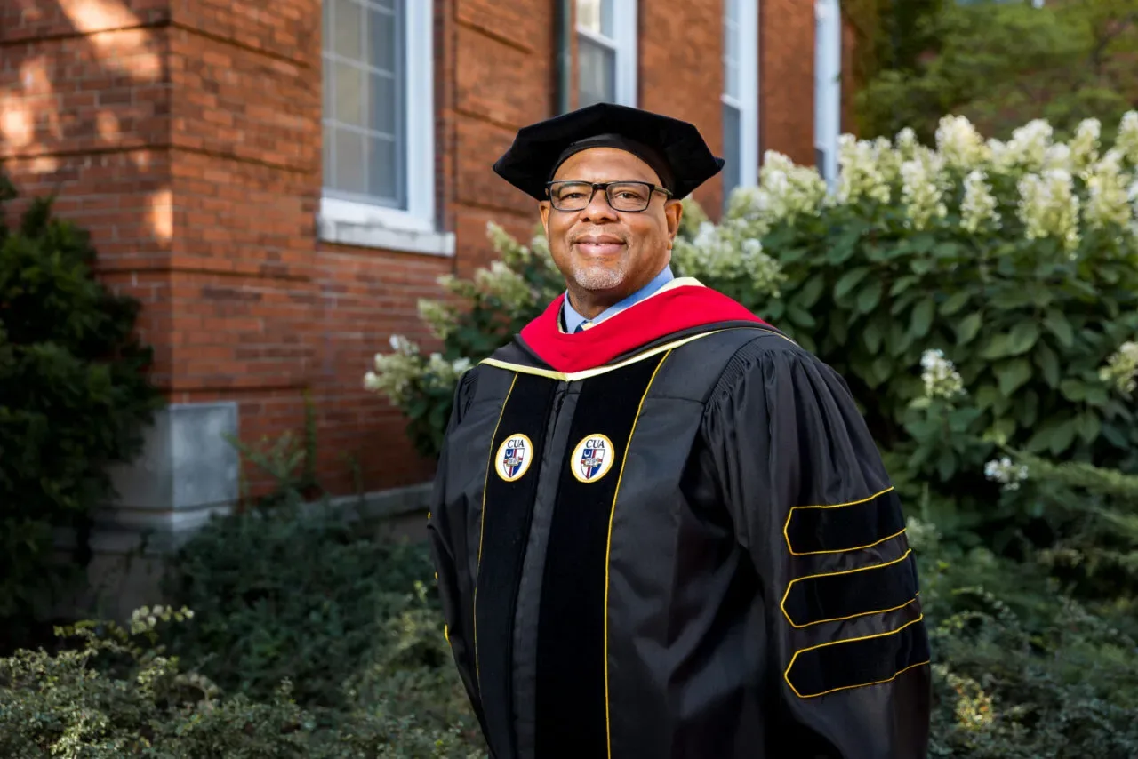 Black scholar in academic doctoral regalia stands in front of red brick building and smiles at the camera