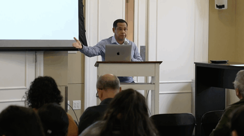 Latino man with short hair lectures from behind a podium to an audience