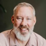 White man with gray-white beard and pink button-up shirt smiles at the camera