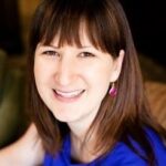 White woman with straight brown hair wearing red dangling earrings and blue shirt smiles at the camera
