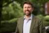 White man with brown hair and beard wearing green blazer and light blue button down shirt smiles at the camera with trees in the background