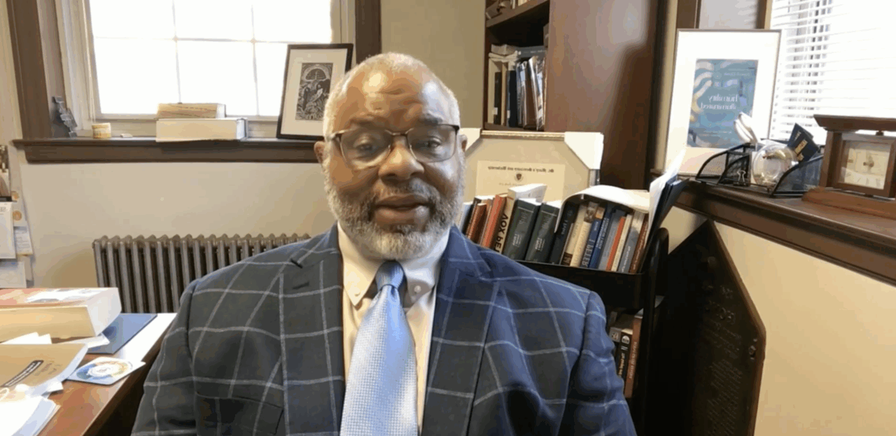 Black professor with balding white hair and beard wearing navy blue windowpane blazer and light blue tie sits in office surrounded by books