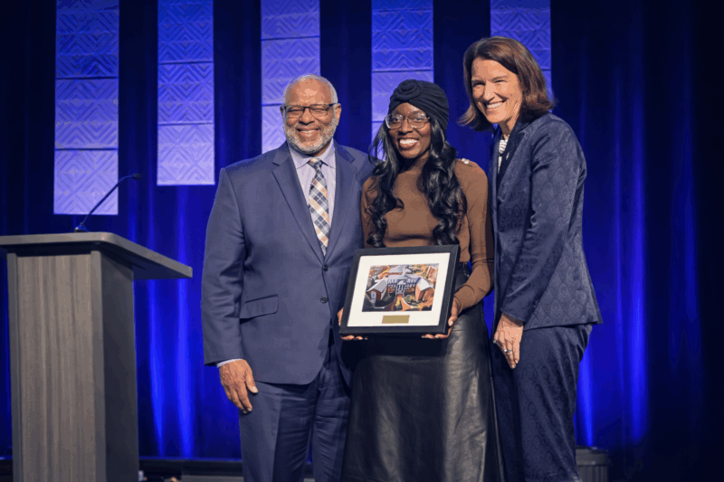 One older black man in blue suit with patterned tie, one middle-aged black woman in brown long sleeve shirt and black turban and tall white lady in navy suit stand on stage together and smile