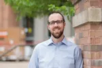 White man with beard and glasses wearing blue button down shirt smiles at the camera while standing beside a brick wall