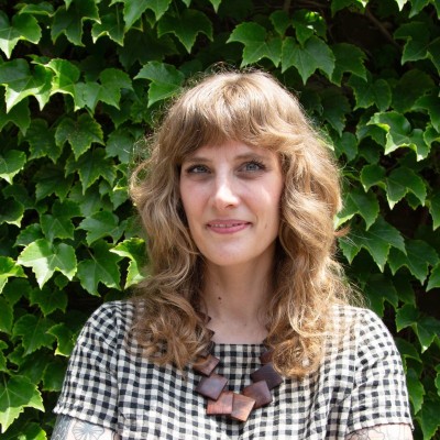Blonde white woman wearing black-and-white check shirt stands in front of ivy-covered wall and smiles at the camera
