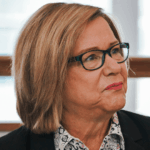 Woman with shoulder-length honey colored hair and dark-rimmed glasses looks past the camera