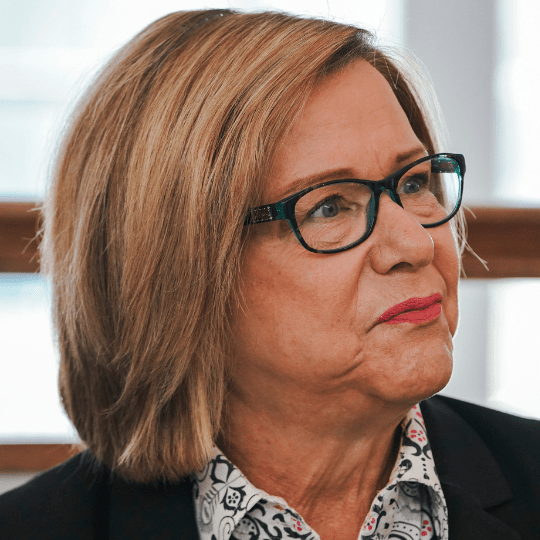 Woman with shoulder-length honey colored hair and dark-rimmed glasses looks past the camera