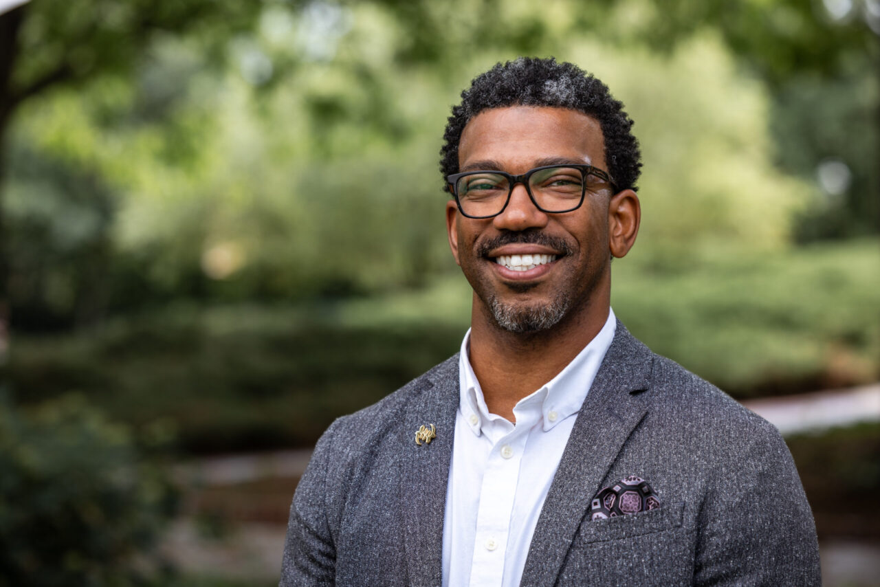 Black man wearing glasses, gray jacket, and white button-down shirt smiles at the camera with trees in the background