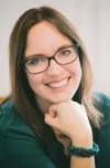 White woman with long straight brown hair and black-rimmed glasses wearing green sweater rests her chin on her fist and smiles at the camera