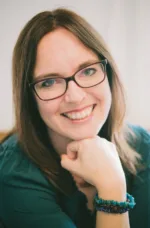 White woman with long straight brown hair and black-rimmed glasses wearing green sweater rests her chin on her fist and smiles at the camera