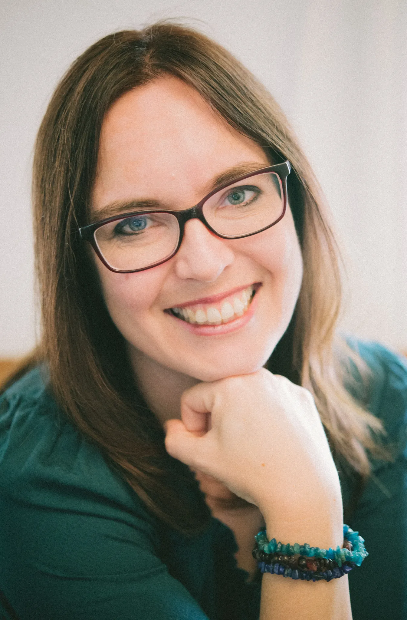 White woman with long straight brown hair and black-rimmed glasses wearing green sweater rests her chin on her fist and smiles at the camera