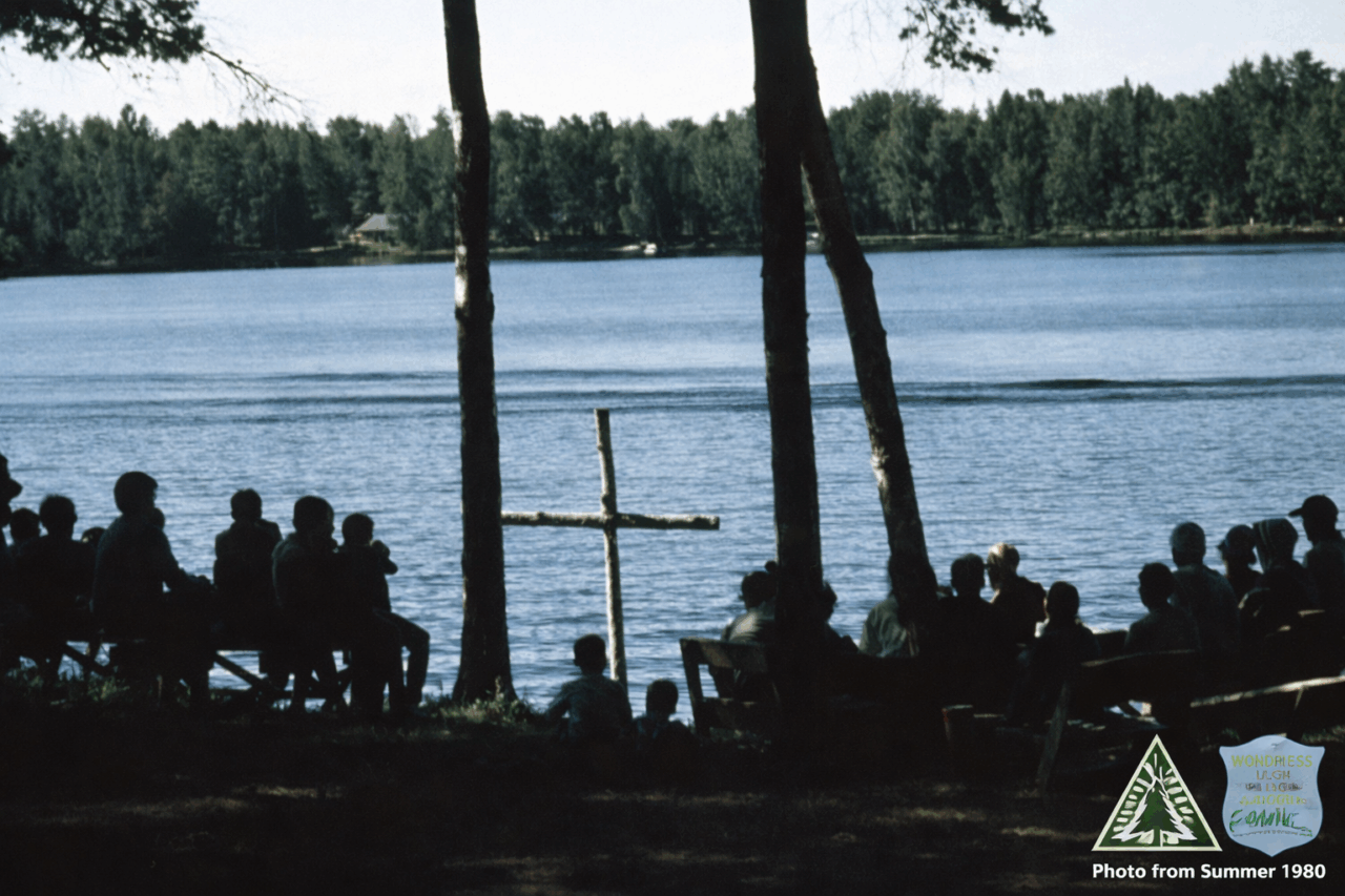 Color photograph showing a lake with tree trunks, a cross, and the silhouettes of campers standing in front of the water