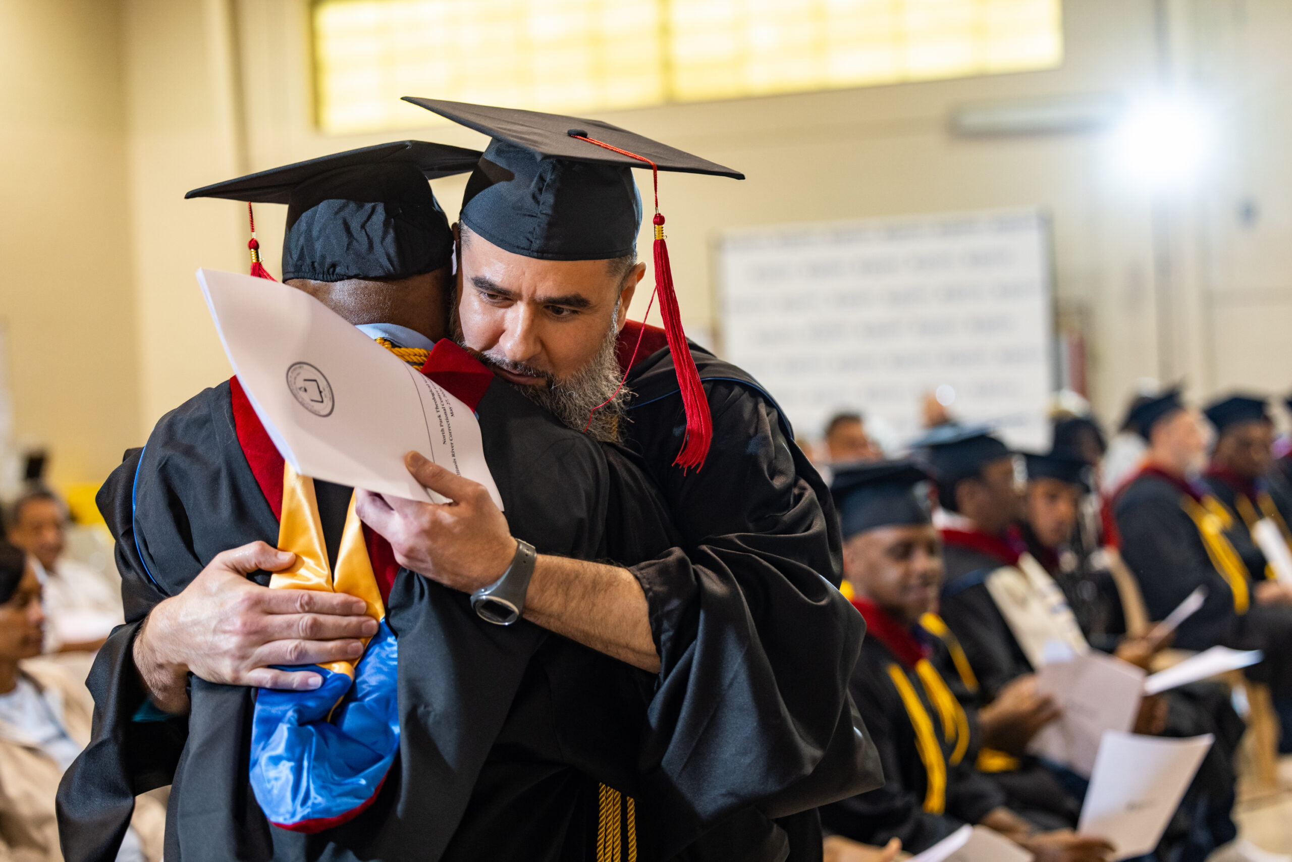 Bearded in man in graduation regalia embraces another man in graduation regalia