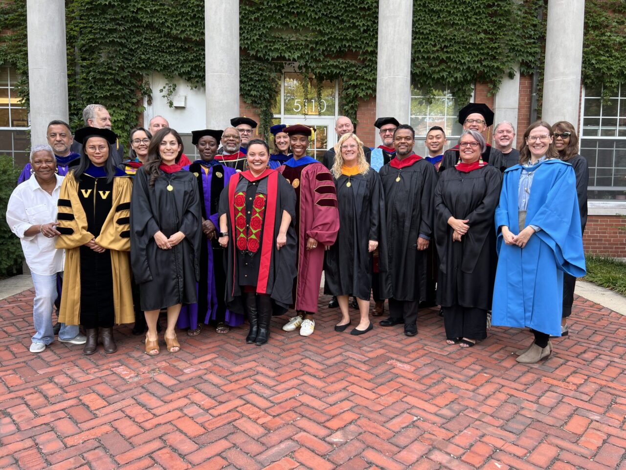 Faculty and staff in academic regalia stand in front of brick building with white columns covered in ivy